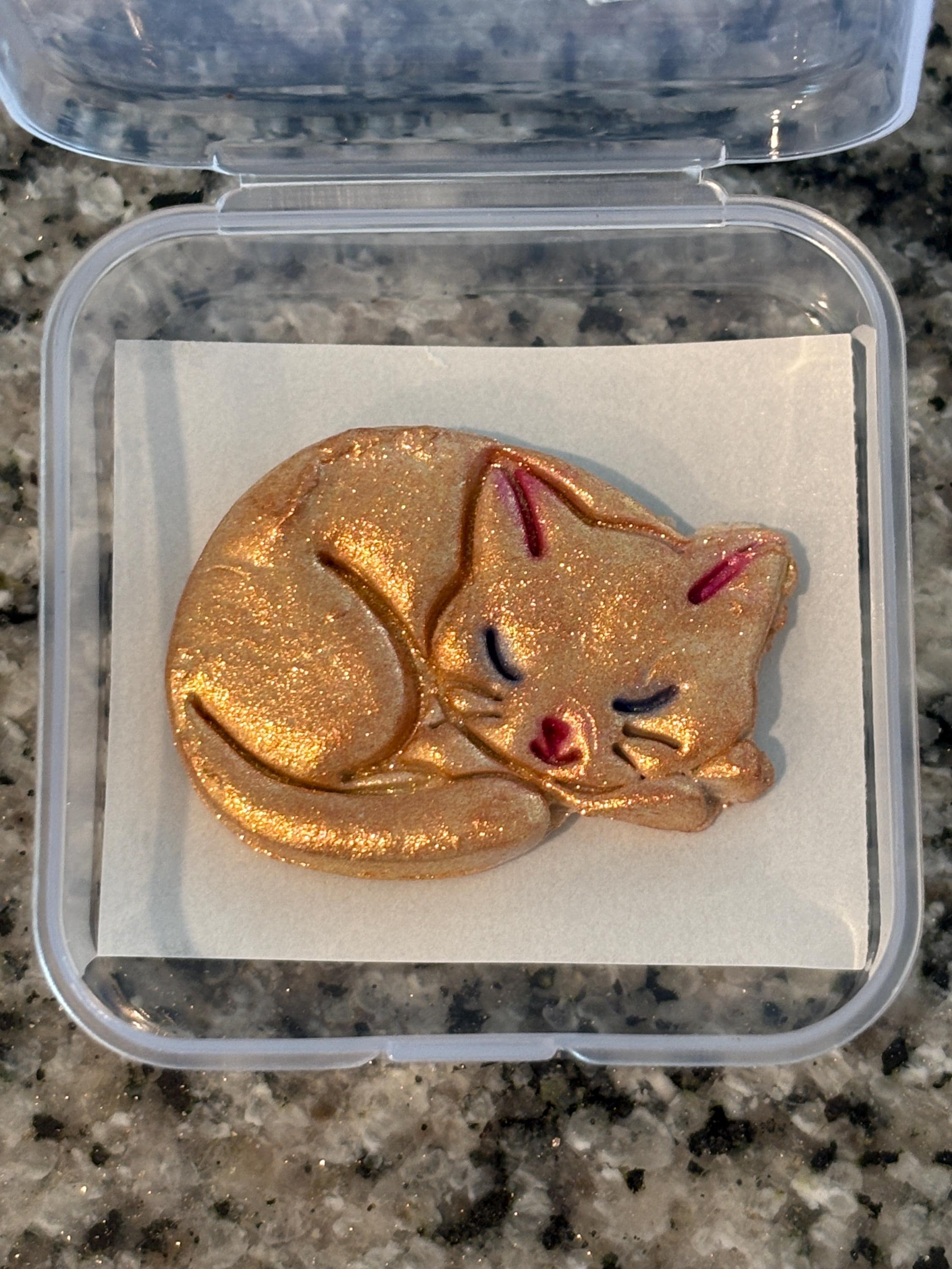 Gold glittery cat-shaped object on a white square inside a clear plastic container.