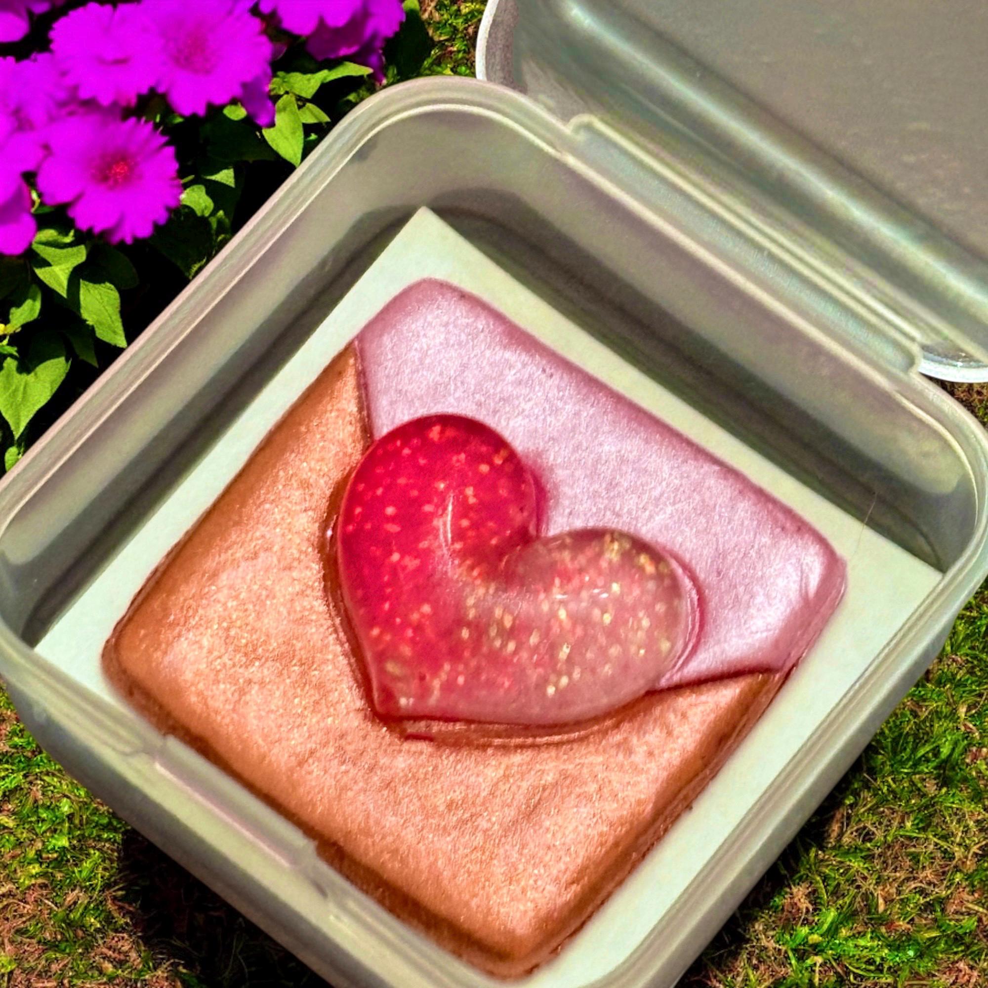 Pink heart-shaped object in a clear container with purple flowers in the background