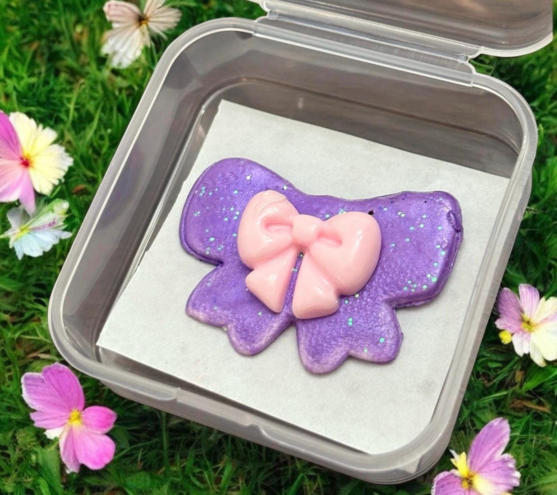 Purple cookie with pink bow in a clear container on grass with flowers