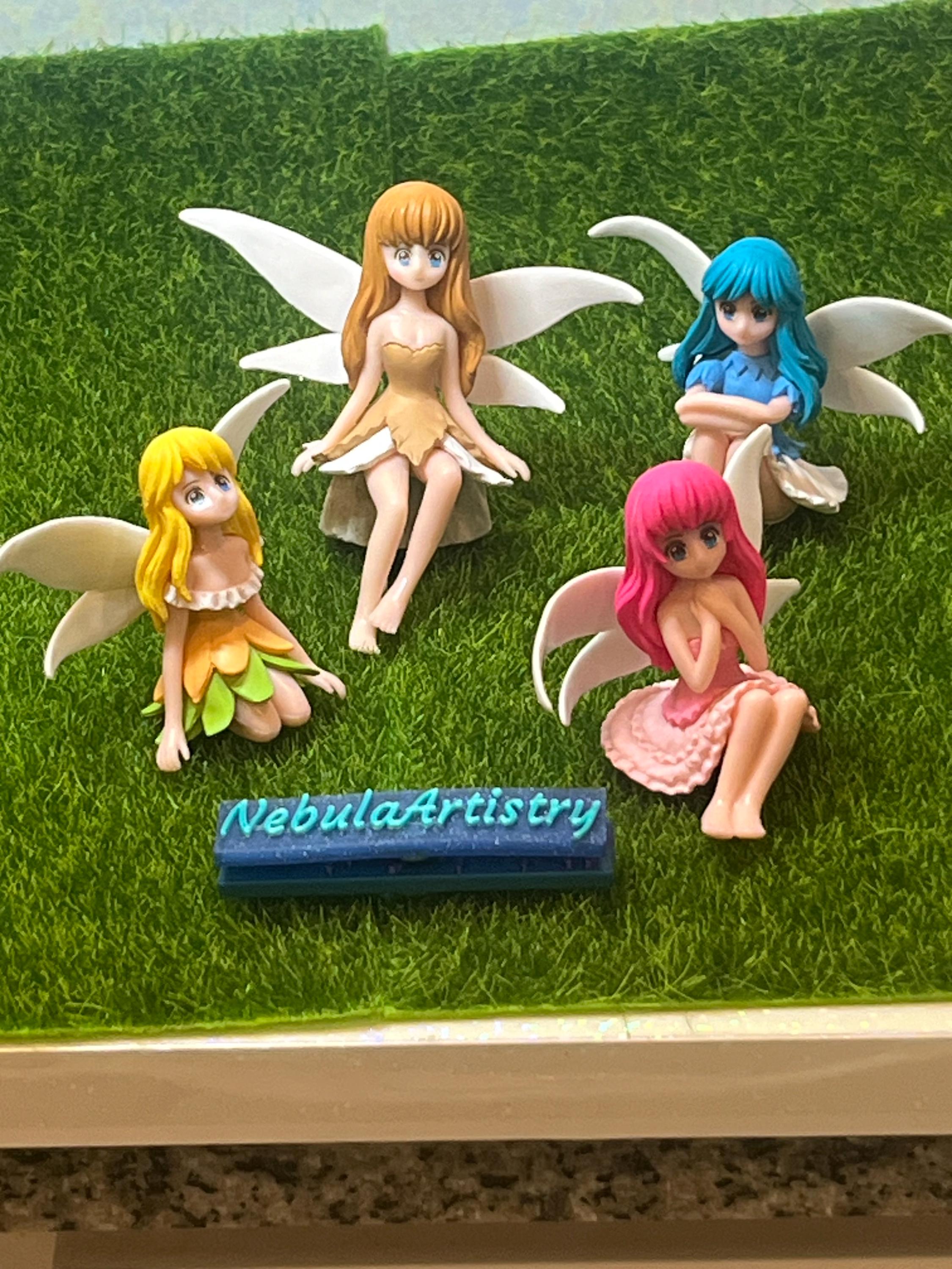 Four fairy figurines with colorful hair on a grass-like surface, branded 'NebulaArtistry'.