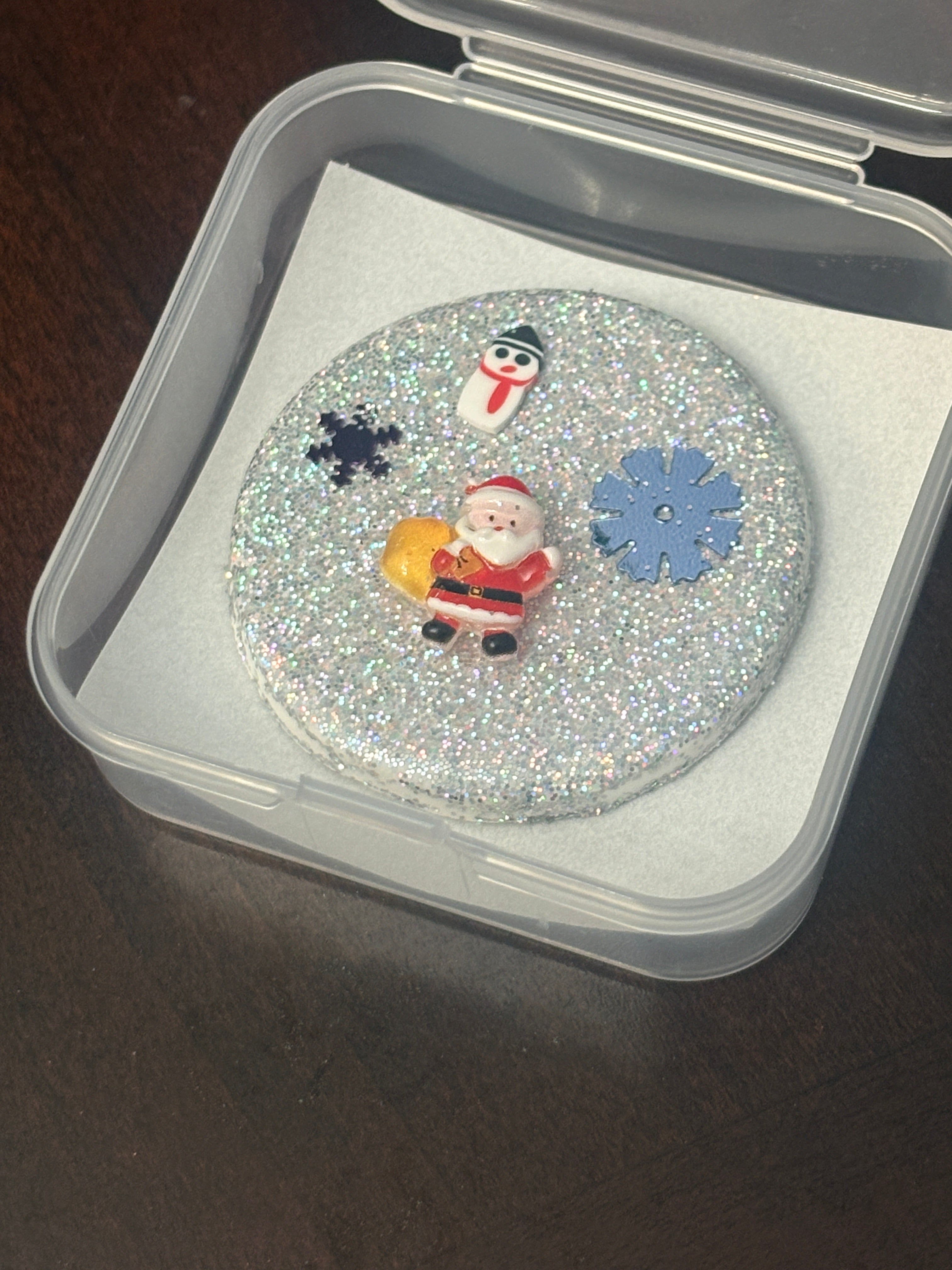 Decorative coaster with snowman and snowflake design on a glittery background, inside a clear plastic container.