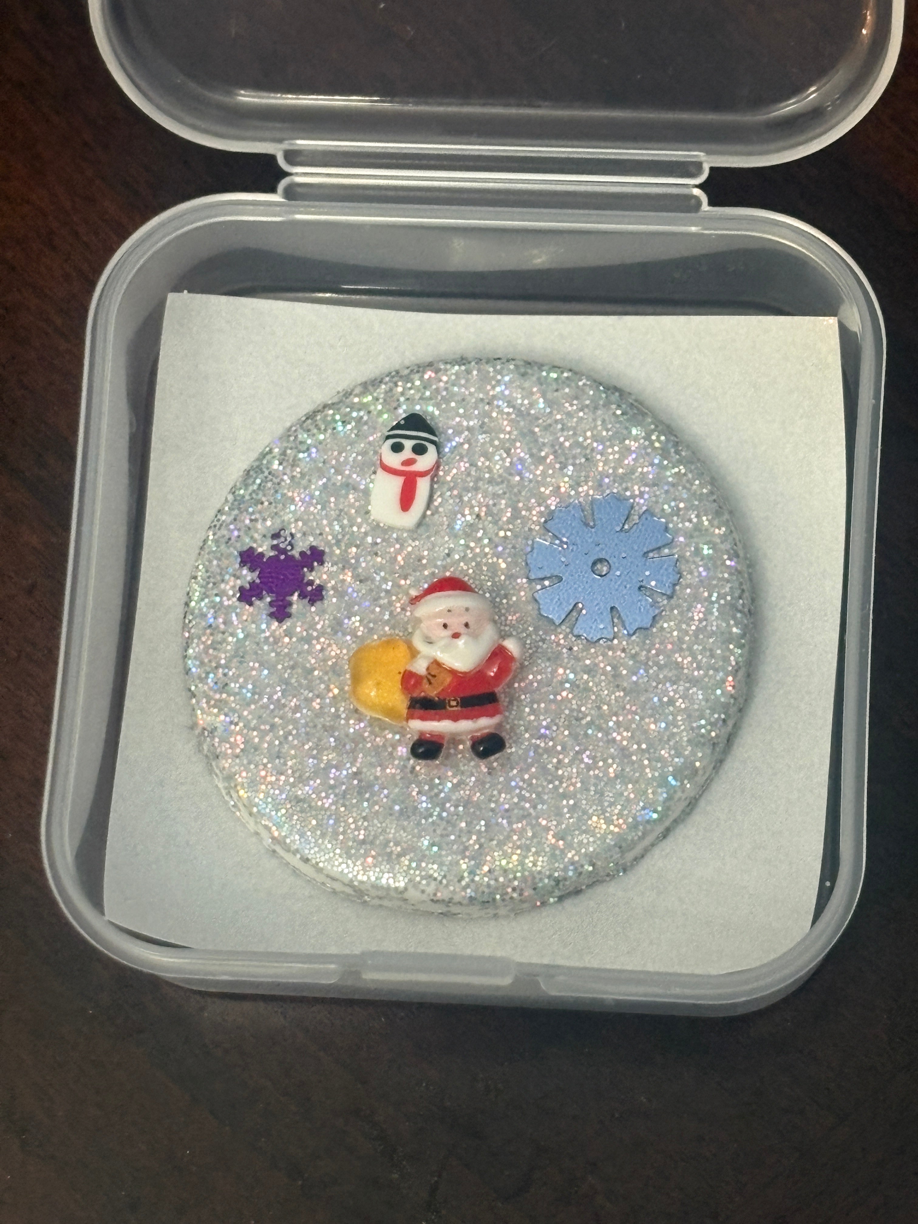 Decorative round object with snowman and snowflake design on a glittery background, inside a clear container.