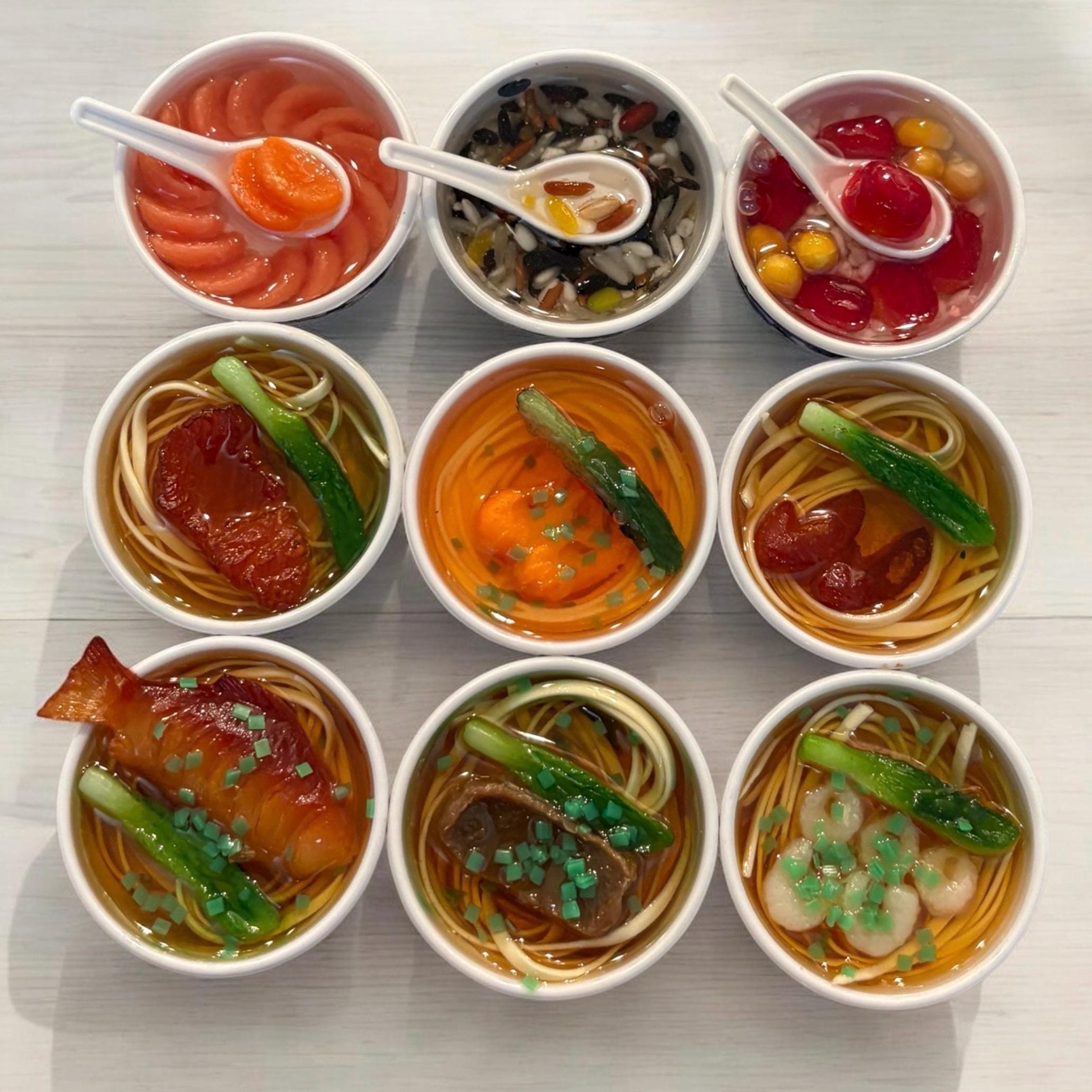 Nine small bowls of various noodle soups with different ingredients on a light wooden surface.