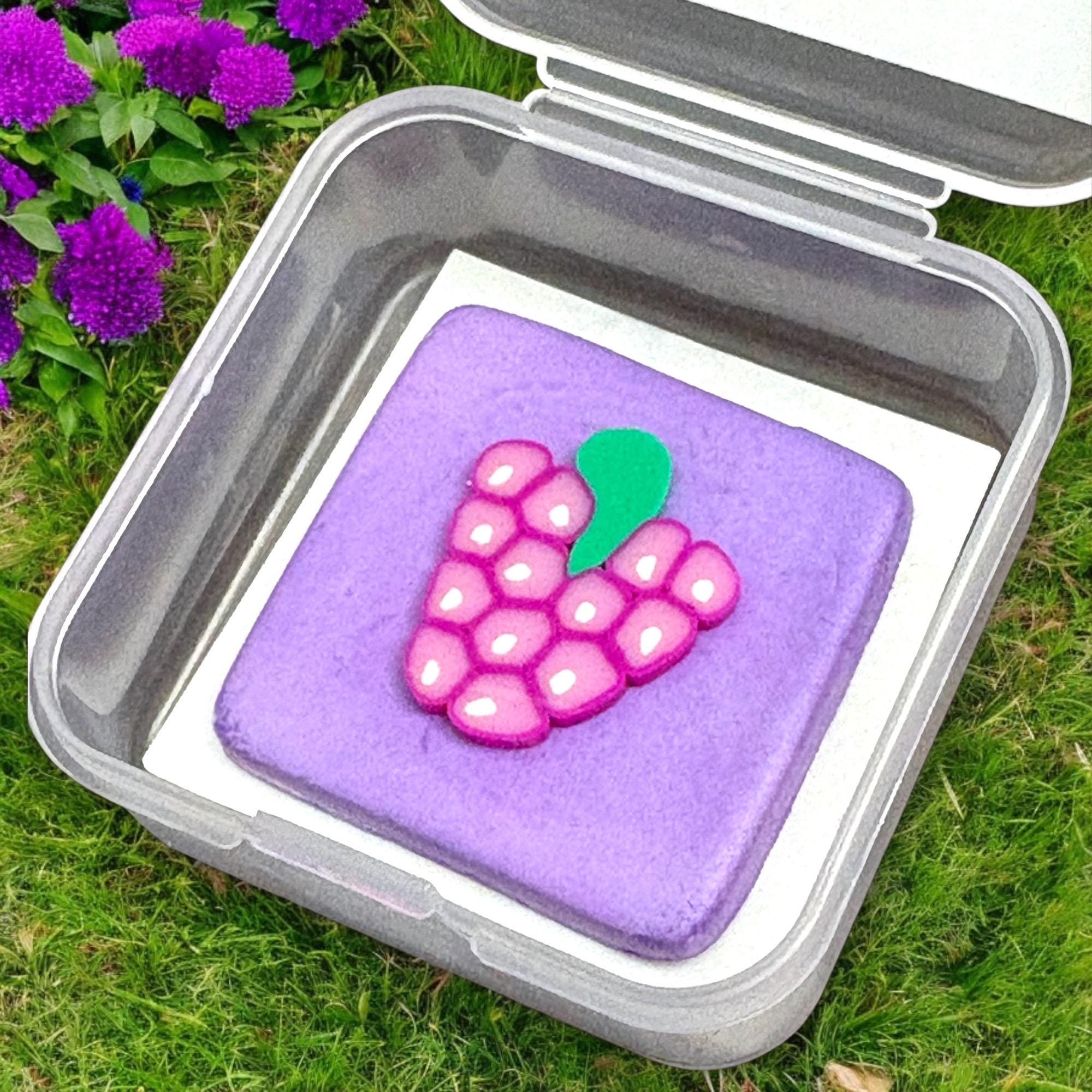 Purple square cake with pink grapes on top in a clear container outdoors.