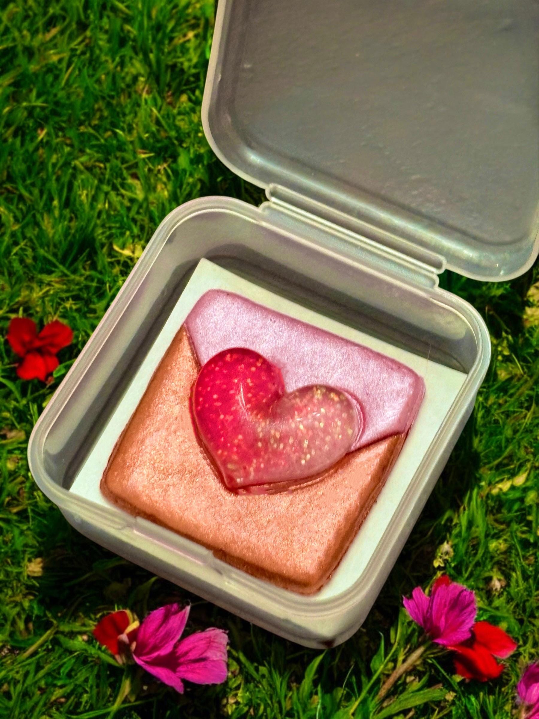 Heart-shaped pink and gold object in a white box on grass with flowers