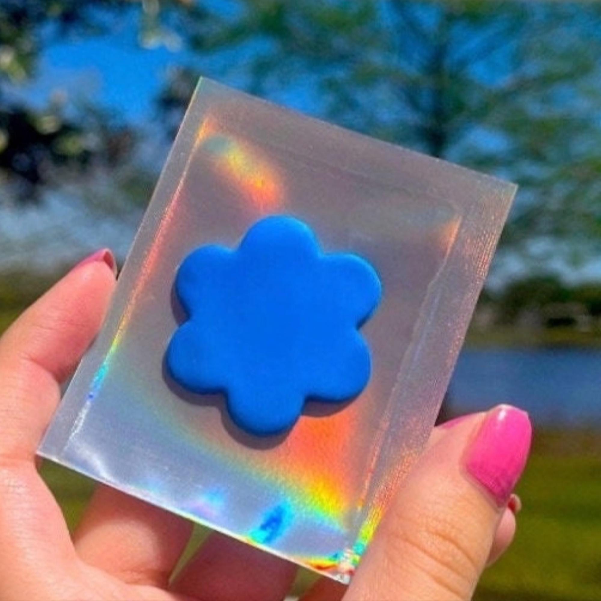 Hand holding a square piece of holographic material with a blue flower shape against a blurred natural background.