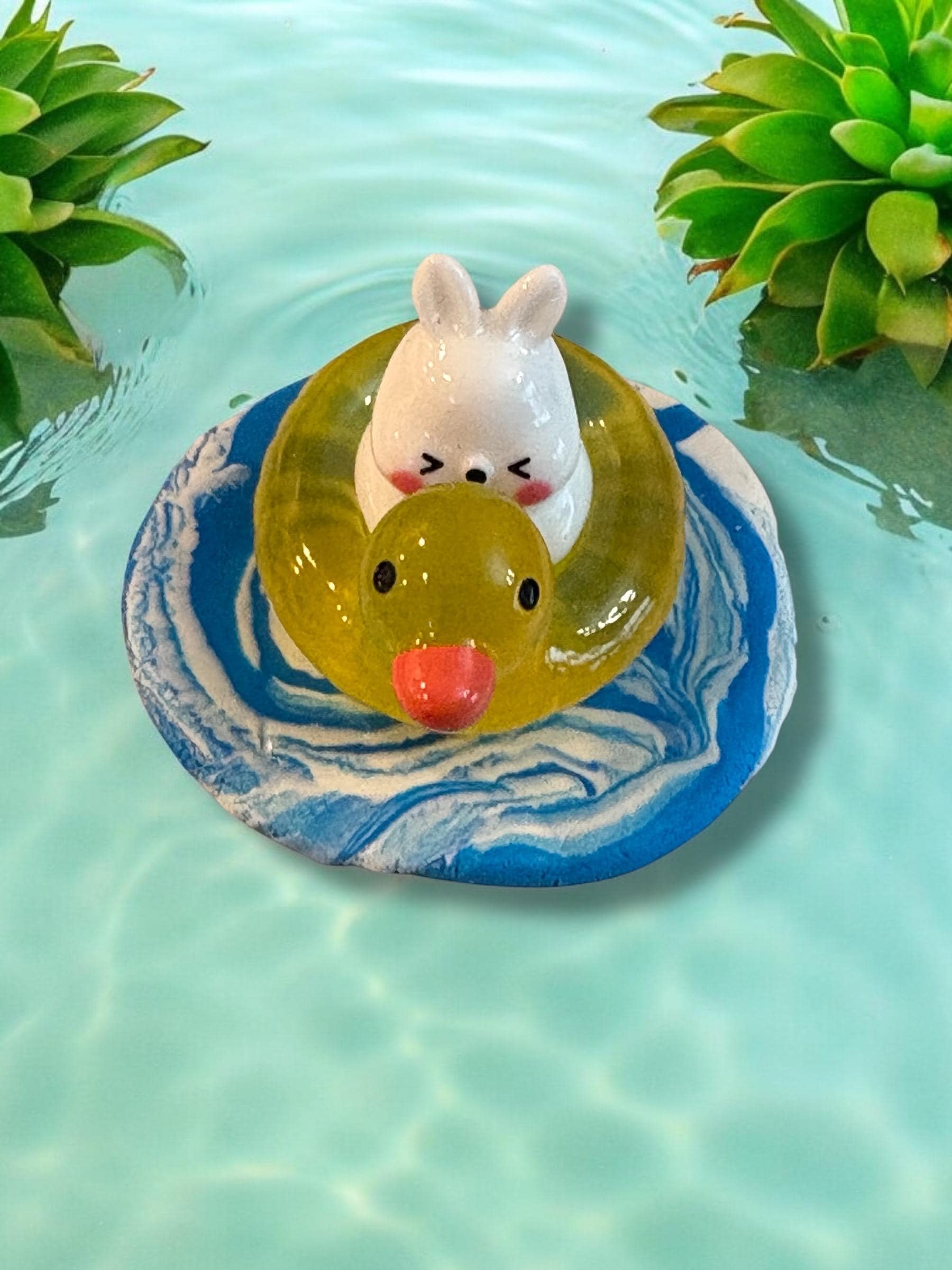 Small rabbit figurine in a yellow rubber duckie on a blue and white shell in clear water with green plants.